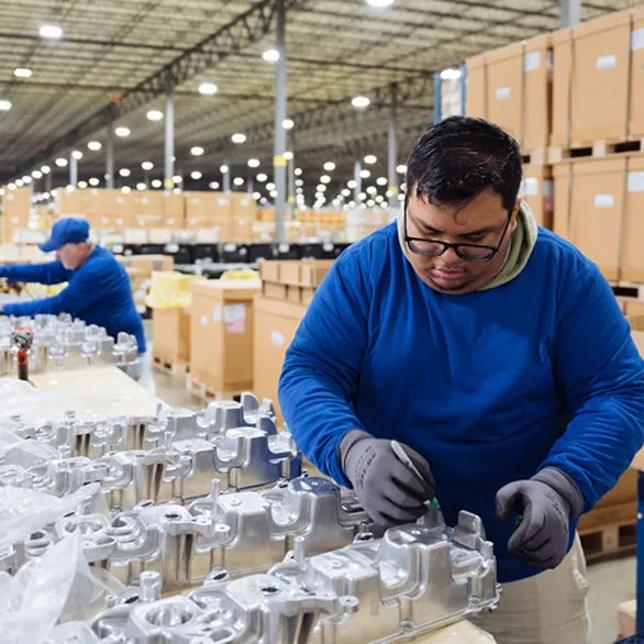 Worker visually inspecting and marking automotive valve covers arranged in rows on a warehouse worktable.