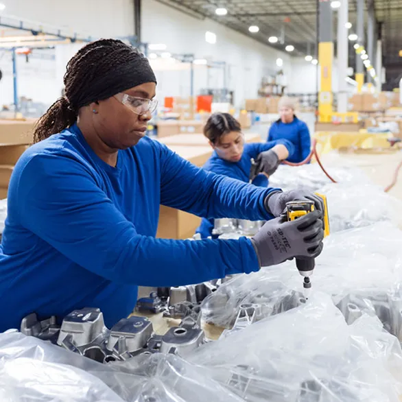 Worker using a handheld power tool to rework threaded holes on an automotive valve cover covered with clear plastic.