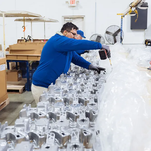 Two workers inspecting and reworking threaded holes on automotive valve covers arranged along a workbench.