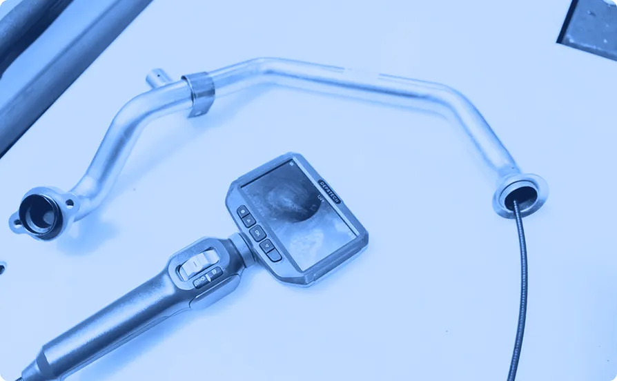 FA metal tube assembly on a work surface is inspected using a digital borescope, with the probe inserted into the tube and the internal surface visible on the handheld screen.