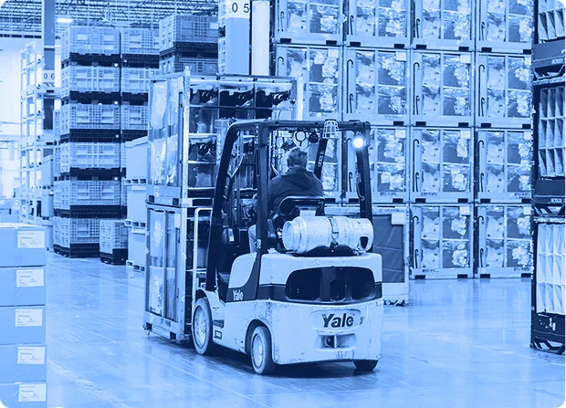 Operator driving a Yale forklift inside a large warehouse filled with stacked shelves and pallets.Forklift operator transporting stacked containers through a warehouse supporting assembly operations.
