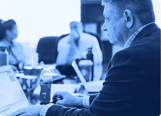 Man focused on his laptop during a business meeting with two blurred colleagues in the background.
