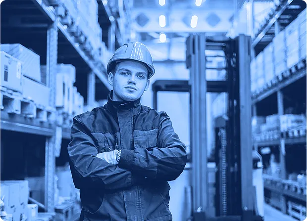 Worker standing in a warehouse aisle with arms crossed near racked inventory and a forklift.