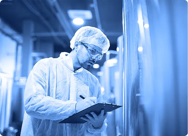 Quality technician wearing protective clothing and writing notes on a clipboard inside a processing area.
