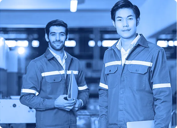 Two employees standing in a manufacturing area holding documents and wearing work uniforms.  