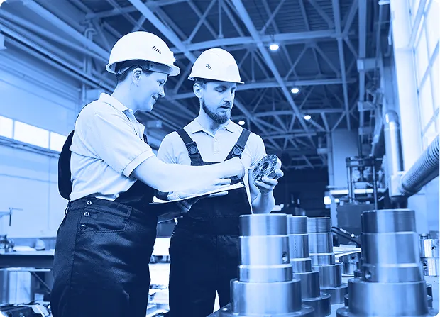Two technicians inspecting cylindrical metal components on a work surface inside a factory.     
