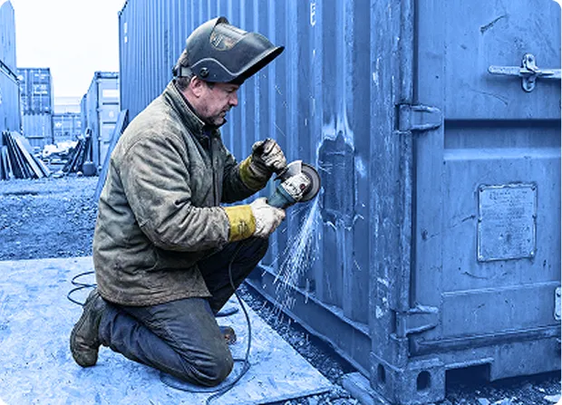 Worker kneeling on a concrete surface while welding the metal door of a large industrial container outdoors.