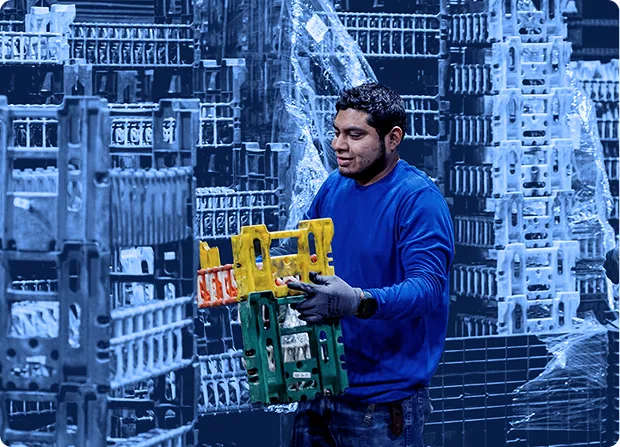 Worker lifting a plastic shipping container from a stacked pallet arrangement inside a warehouse with wrapped pallets in the background.