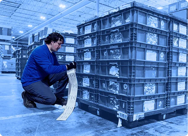 Worker kneeling beside stacked red plastic containers while reading a printed label inside a warehouse.