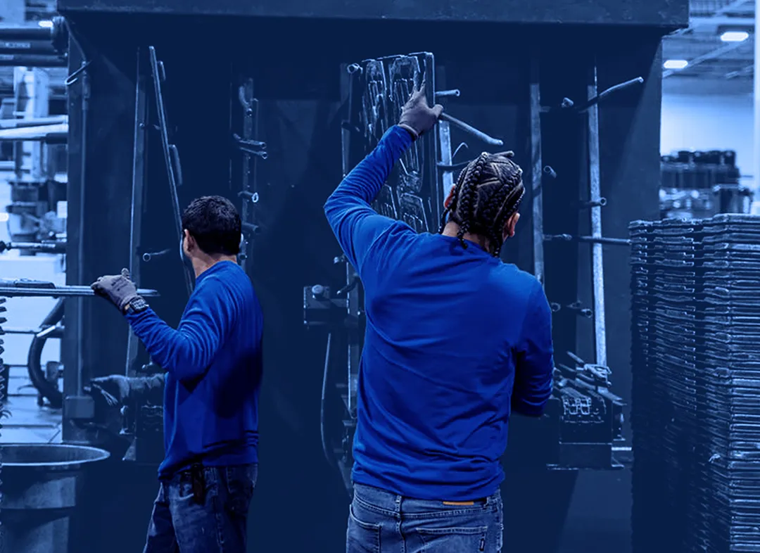 Two workers wearing gloves load and remove flat metal parts from a large blue industrial pressure washing machine.