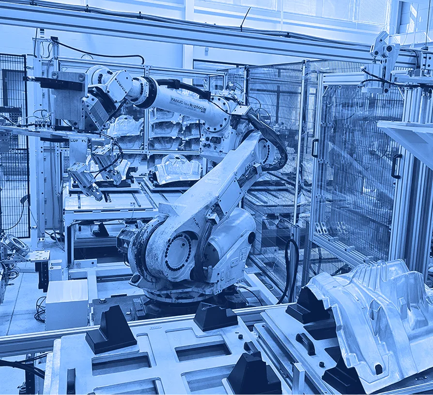 Industrial robotic arms operate within an automated manufacturing cell, positioning metal components on fixtures along a conveyor inside a factory.