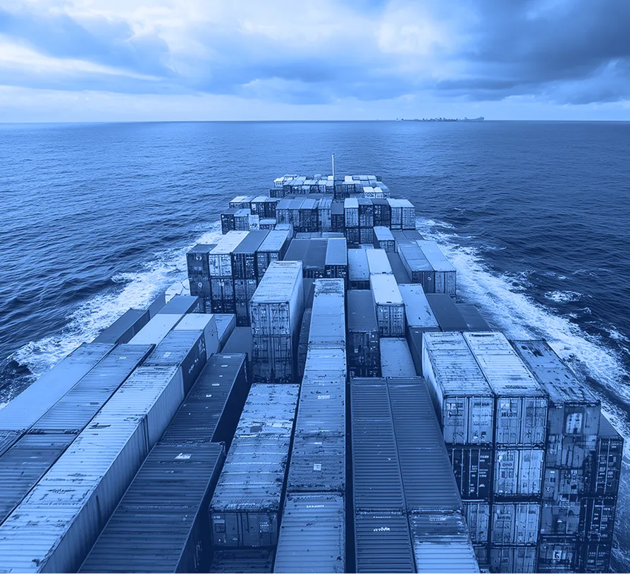 View from the stern of a cargo ship showing rows of stacked shipping containers moving through open ocean under a cloudy sky.