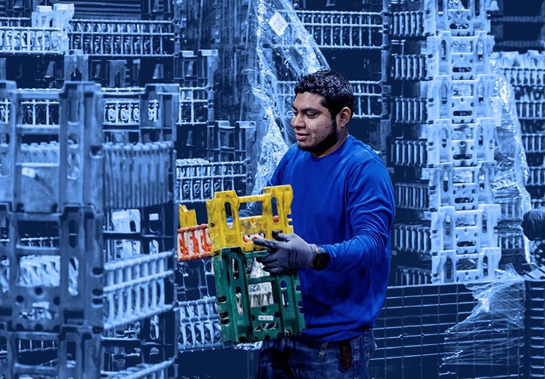 Worker lifting a plastic shipping container from a stacked pallet arrangement inside a warehouse with wrapped pallets in the background.