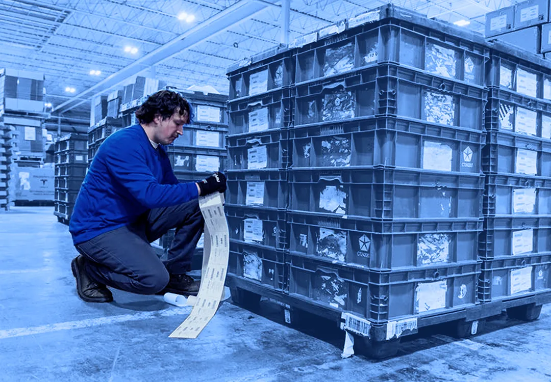 Worker kneeling beside stacked red plastic containers while reading a printed label inside a warehouse.  