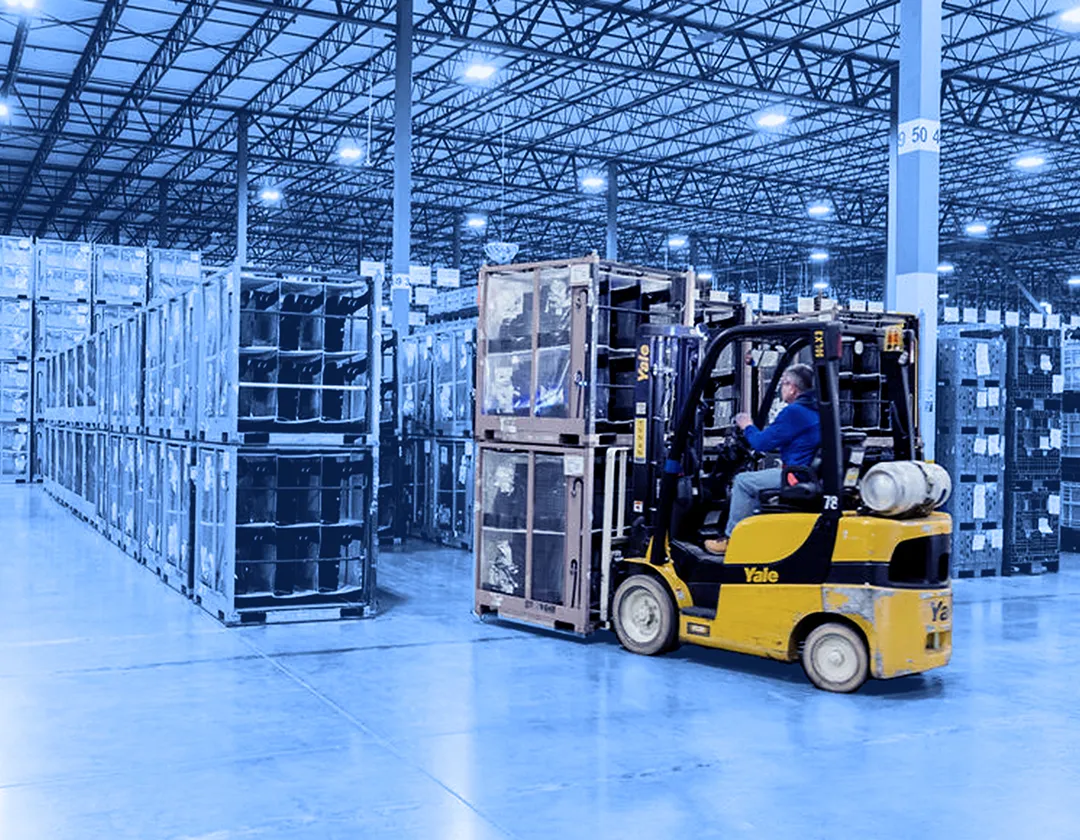 Worker operating a forklift inside a large warehouse, moving stacked metal containers and plastic bins arranged in rows on a concrete floor.  