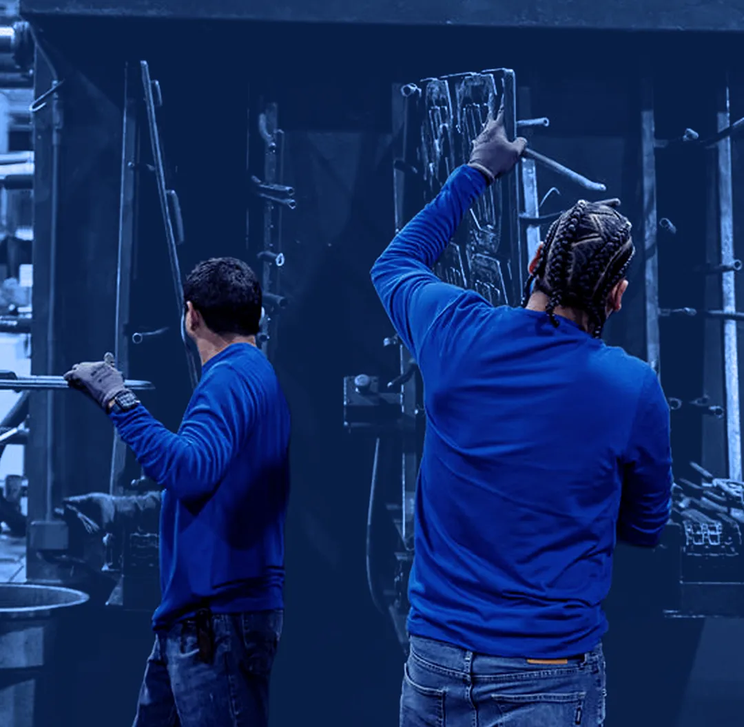 Two workers wearing gloves load and remove flat metal parts from a large blue industrial pressure washing machine.