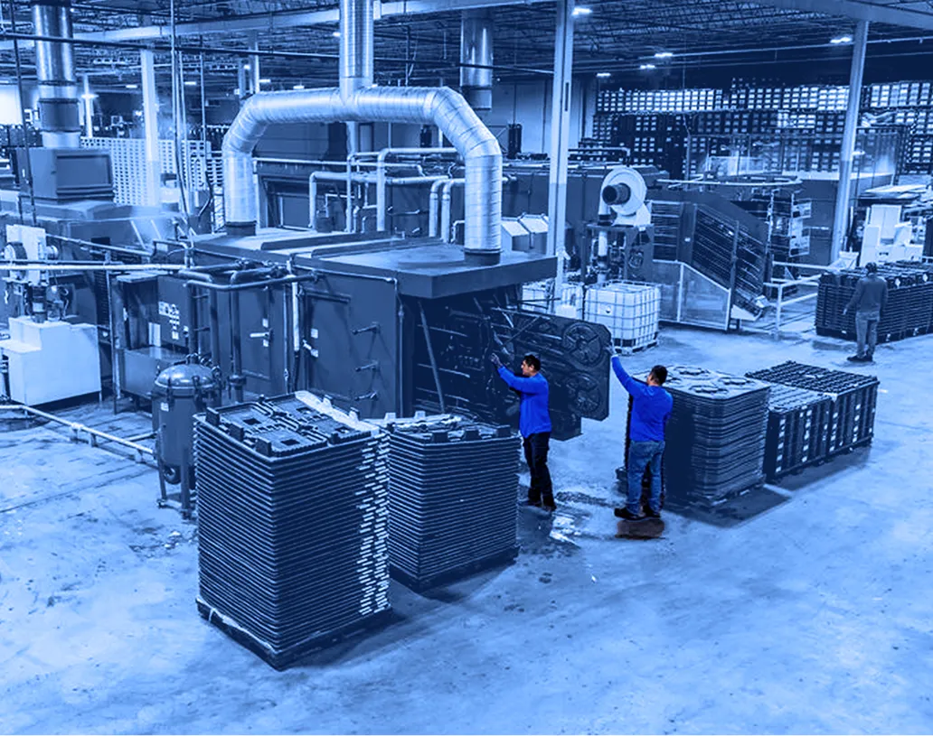 Wide view of a factory floor showing several workers handling stacks of flat plastic pallets around large blue industrial processing machines with overhead ducting.