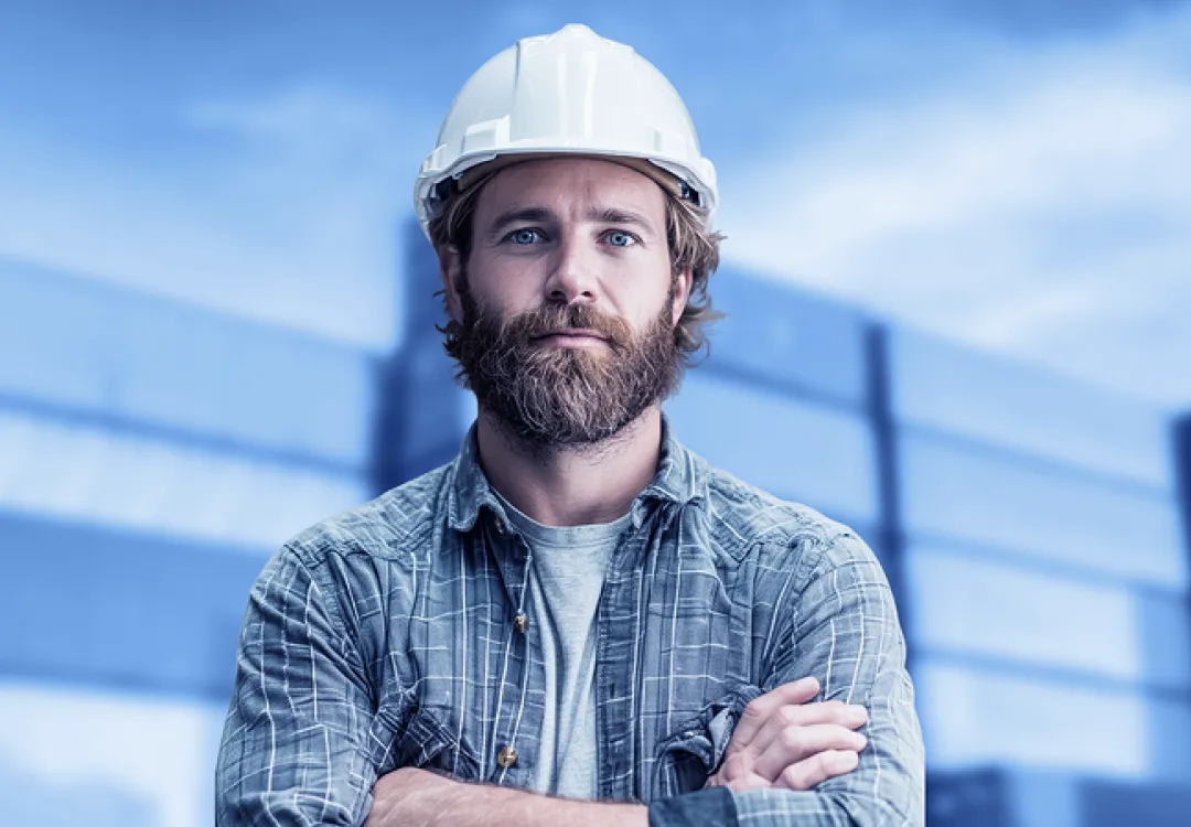 Man wearing a white hard hat stands outdoors with arms crossed, facing the camera with an industrial building blurred in the background.
