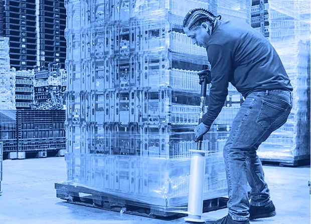 Worker wearing gloves uses a handheld stretch wrap tool to secure stacked plastic containers on a pallet inside a warehouse.  
