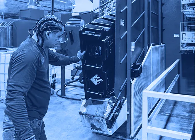 Worker wearing gloves positions a black plastic container into a large blue industrial washing machine inside a factory.