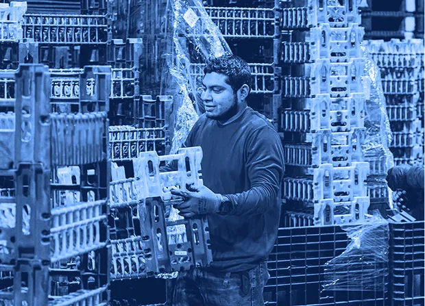Worker lifting a plastic shipping container from a stacked pallet arrangement inside a warehouse with wrapped pallets in the background.       