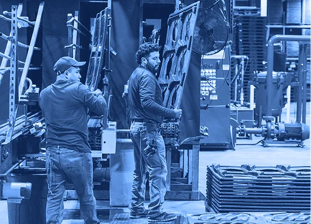 Two workers wearing gloves remove molded plastic panels from a large blue industrial washing machine on a factory floor, with stacked panels nearby.  