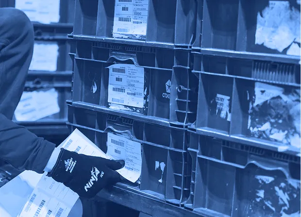 Worker wearing gloves places labeled items into red plastic storage bins arranged on shelving.
