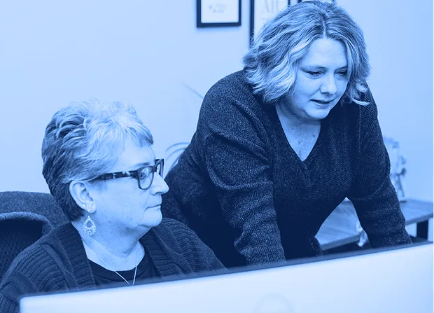 Two office employees review information on a desktop computer at a shared workstation in an office setting.  