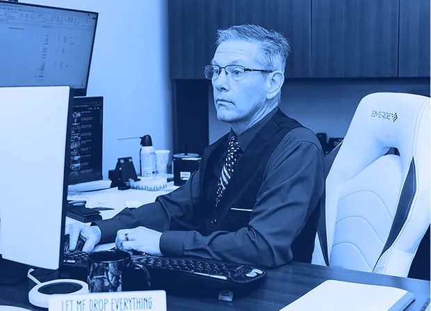 Office employee seated at a desk works on a computer workstation with dual monitors, keyboard, and paperwork in a private office.  
