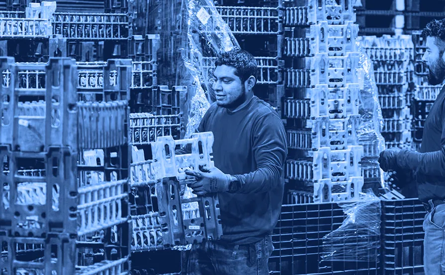 Worker lifting a plastic shipping container from a stacked pallet arrangement inside a warehouse with wrapped pallets in the background.