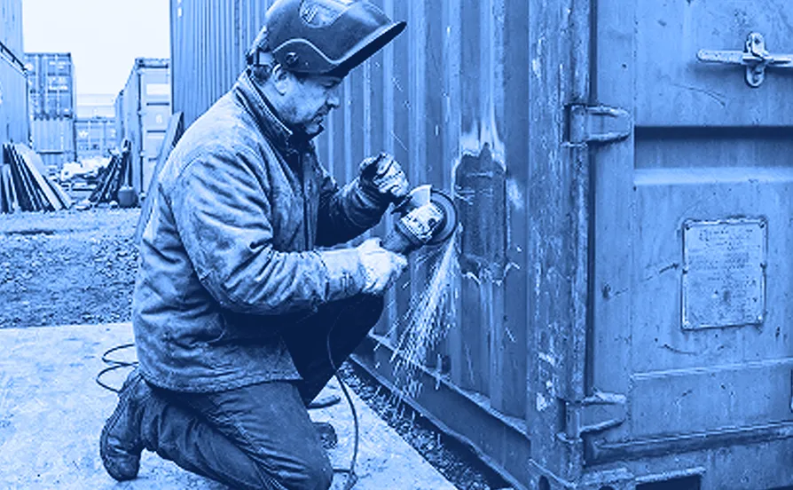 Worker kneeling on a concrete surface while welding the metal door of a large industrial container outdoors.