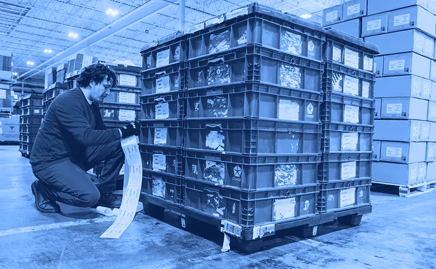 Worker kneeling beside stacked red plastic containers while reading a printed label inside a warehouse.