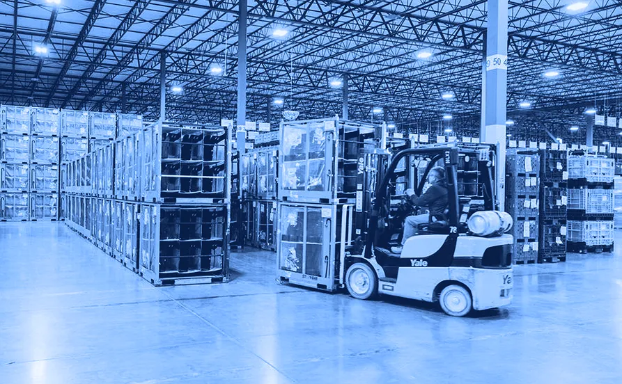 Worker operating a forklift inside a large warehouse, moving stacked metal containers and plastic bins arranged in rows on a concrete floor.