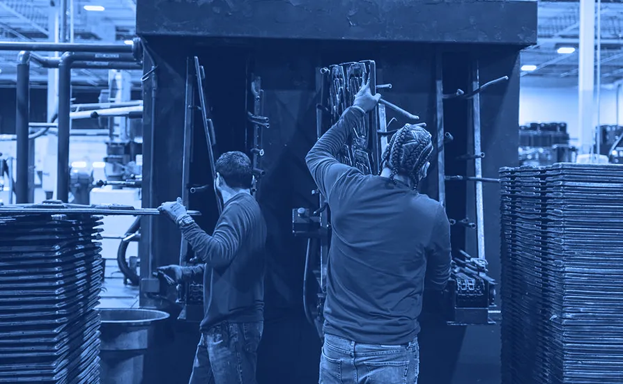 Two workers wearing gloves load and remove flat metal parts from a large blue industrial pressure washing machine.