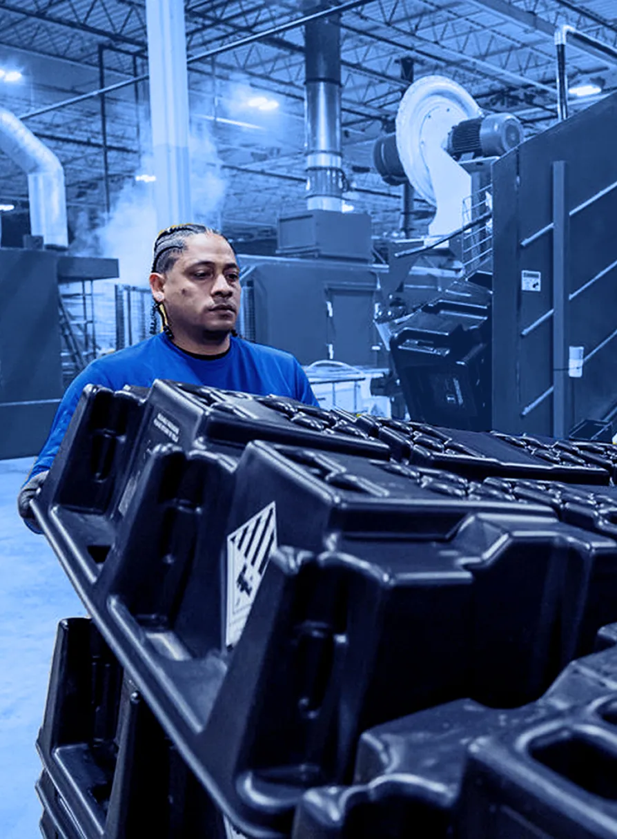 Worker carries stacked black plastic containers inside a factory with large blue industrial washing machines in the background.
