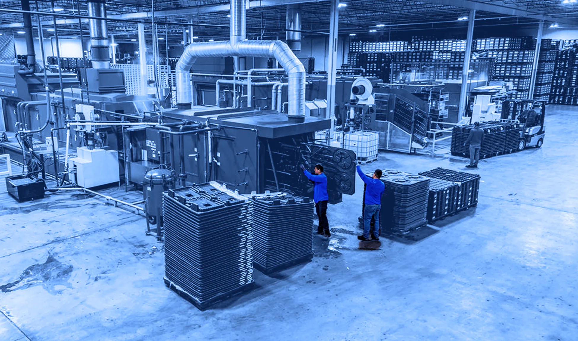Wide view of a factory floor showing workers handling stacks of flat plastic pallets around large blue industrial processing machines with overhead ducting.