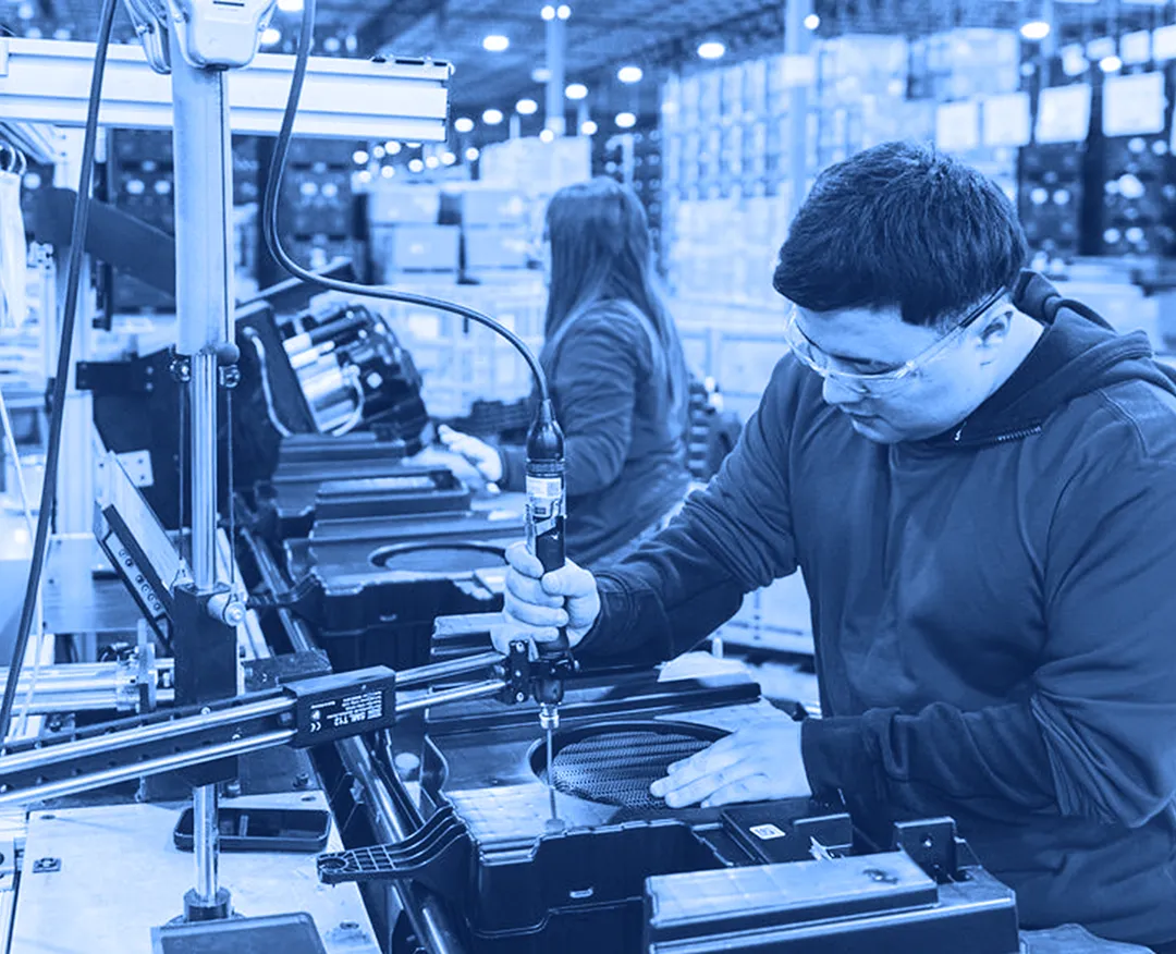 Worker using a handheld power tool to fasten a component into a plastic housing at an assembly station inside a warehouse.