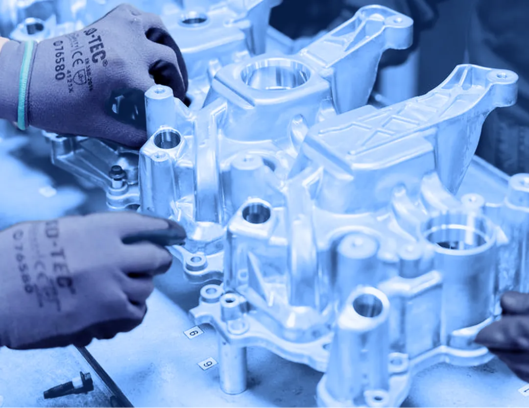 Two workers wearing gloves positioning aluminum cast components on a metal work surface with fasteners arranged nearby.