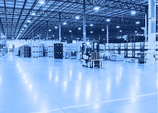Forklift operator driving through a warehouse aisle with pallet racking, stacked boxes, and open floor space visible.