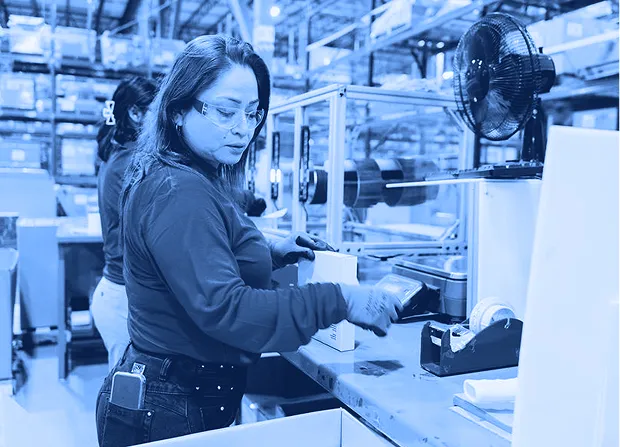 A worker wearing safety glasses and gloves scans a small box at a packing station inside a warehouse with shelving and equipment in the background.