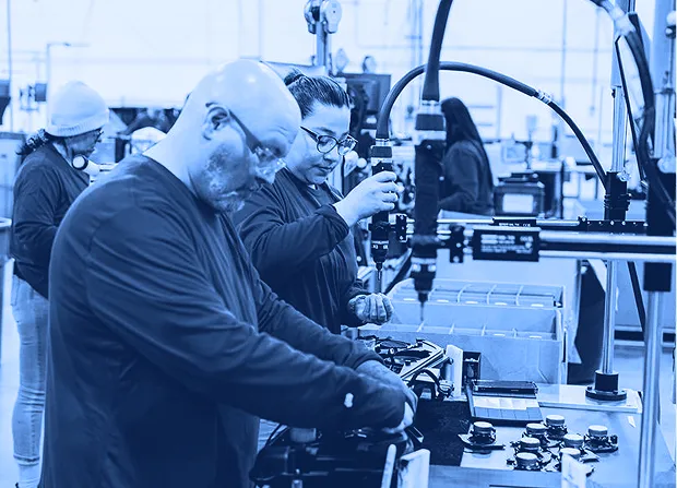 Two workers wearing safety glasses operating handheld power tools at an assembly workstation with parts, fixtures, and boxes visible around them.
