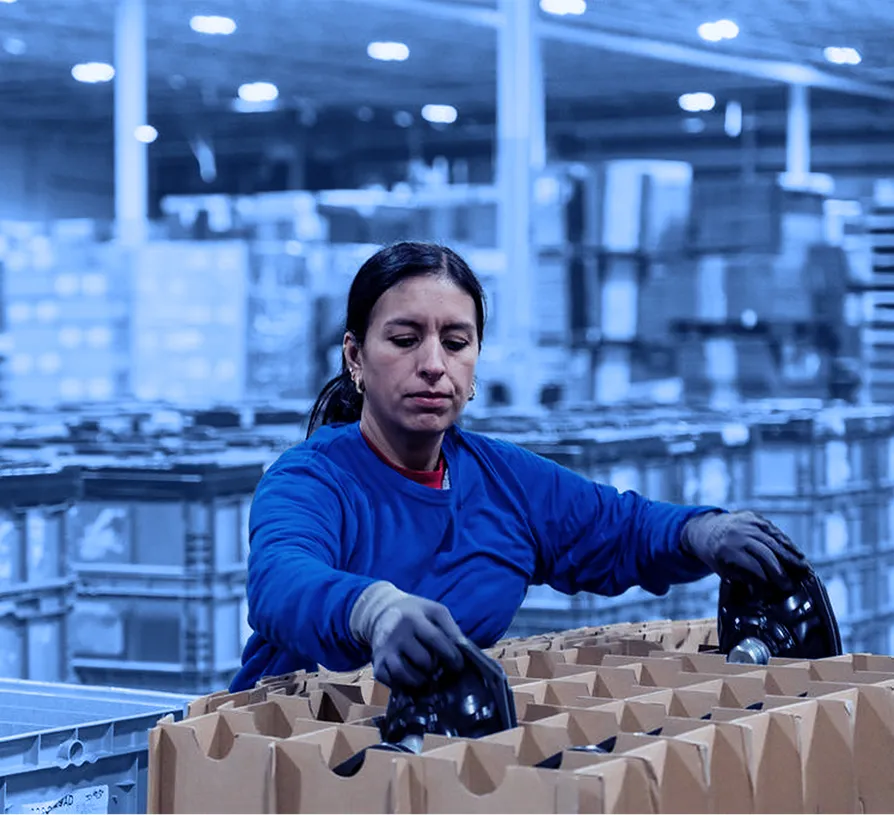 Worker wearing gloves places black plastic components into a cardboard tray on a warehouse floor with stacked containers in the background.