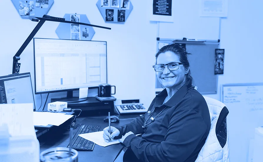Office worker wearing glasses seated at a desk writing on paper with a computer monitor, keyboard, and bulletin boards visible in the workspace.