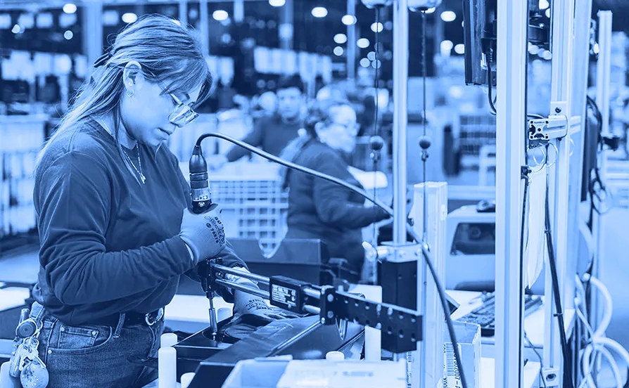 Worker wearing gloves and safety glasses using a handheld power tool to fasten a component at an assembly station inside a warehouse.