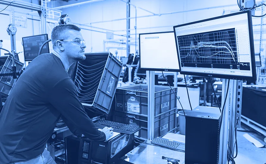 Worker wearing safety glasses operating a computer at a factory testing station while viewing line graphs on two monitors.