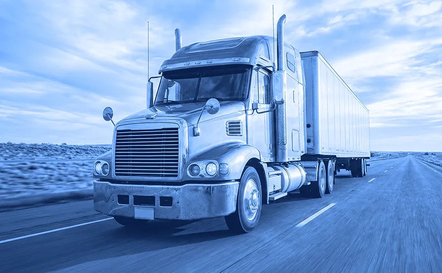 Semi truck pulling a box trailer driving along a paved highway through an open landscape under a cloudy sky.