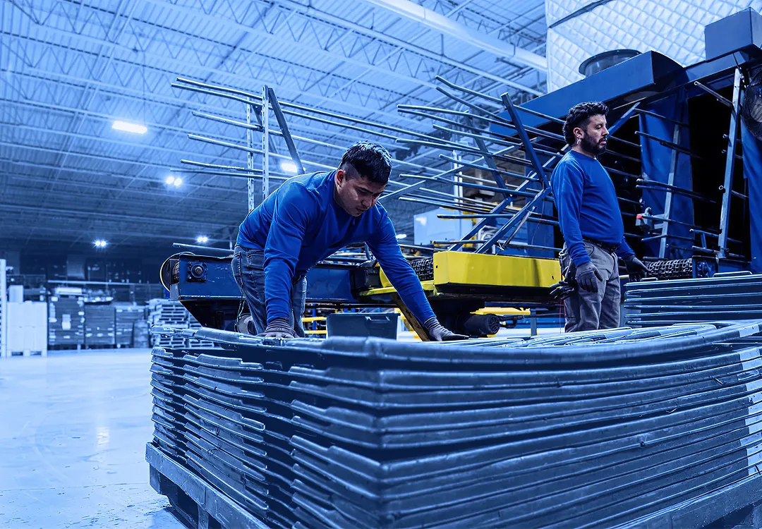 Worker in blue shirt stacks black plastic returnable trays on a pallet in a warehouse while another worker stands nearby beside a large high-pressure washer.