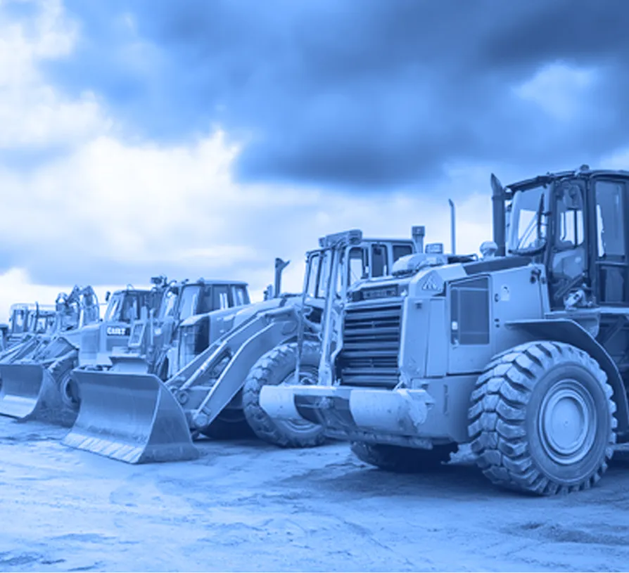 Row of large yellow wheel loaders with front buckets parked on a dirt surface under an overcast sky. 