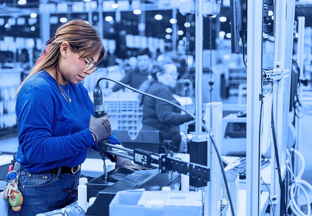 Worker wearing gloves and safety glasses using a handheld power tool to fasten a component at an assembly station inside a warehouse.