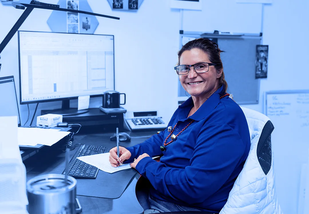 Office worker wearing glasses seated at a desk writing on paper with a computer monitor, keyboard, and bulletin boards visible in the workspace.